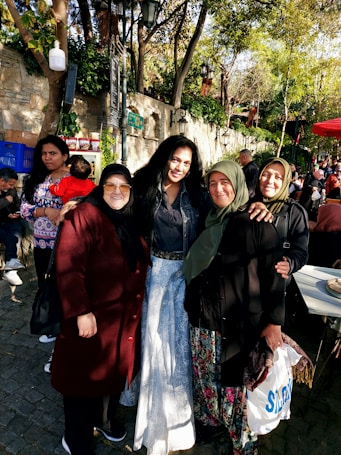 A group of women standing together outdoors, surrounded by trees and people in a lively environment. Some are wearing headscarves and smiling, while others are engaged in conversations. Various people and elements like trees, a stone wall, and a red canopy add depth to the setting.