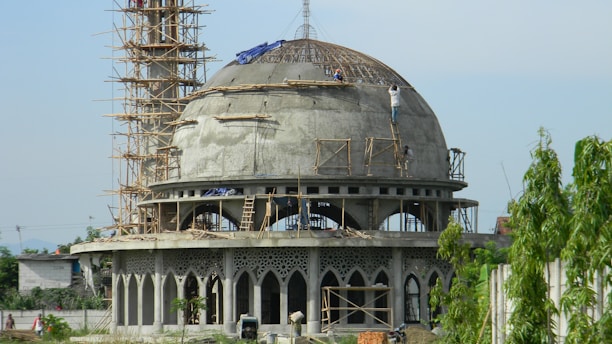 A large dome-shaped building under construction, surrounded by scaffolding and workers. The structure features intricate arched windows and decorative elements. Scattered construction materials and greenery are visible in the foreground.