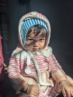 A young child wearing a striped winter outfit with a hood and a matching knitted cap. The child is indoors, illuminated by natural light that highlights their serious expression. The sweater features a combination of white, pink, and blue stripes.