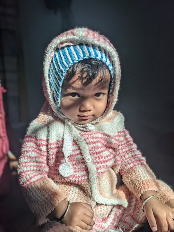 A young child wearing a striped winter outfit with a hood and a matching knitted cap. The child is indoors, illuminated by natural light that highlights their serious expression. The sweater features a combination of white, pink, and blue stripes.