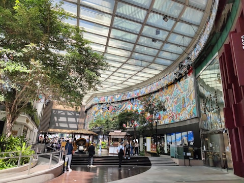 A spacious, modern indoor area with a high glass ceiling allowing natural light to enter. The walls are adorned with colorful, abstract murals featuring various patterns and shapes. Lush green trees and plants add a touch of nature to the space. People are walking around, some gathered near a small kiosk or stall.