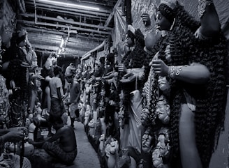 A narrow corridor filled with large, intricately crafted statues, likely depicting mythological figures, lined up against the walls. Several artisans are working meticulously on the statues, which have detailed features and ornate garments. The atmosphere is busy with artisans focusing on their craft.