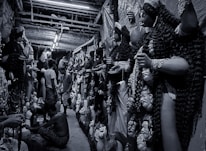 A narrow corridor filled with large, intricately crafted statues, likely depicting mythological figures, lined up against the walls. Several artisans are working meticulously on the statues, which have detailed features and ornate garments. The atmosphere is busy with artisans focusing on their craft.