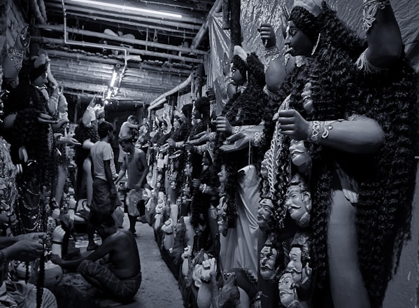 A narrow corridor filled with large, intricately crafted statues, likely depicting mythological figures, lined up against the walls. Several artisans are working meticulously on the statues, which have detailed features and ornate garments. The atmosphere is busy with artisans focusing on their craft.