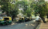 A tree-lined street features several green and yellow auto rickshaws parked alongside the curb. A couple of pedestrians walk nearby, while sunlight filters through the leaves, casting dappled shadows on the road. A white car is also parked further down the street.