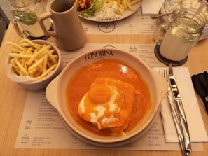 A plate with a dish covered in a bright orange sauce is placed on a table alongside a bowl of French fries. A glass jar of lemonade with a mint leaf, a ceramic jug, and a knife and fork wrapped in a napkin are also present. A menu can be seen on the table.