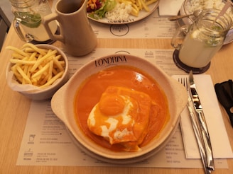 A plate with a dish covered in a bright orange sauce is placed on a table alongside a bowl of French fries. A glass jar of lemonade with a mint leaf, a ceramic jug, and a knife and fork wrapped in a napkin are also present. A menu can be seen on the table.