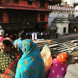 A group of people dressed in colorful traditional clothing gather near a stone platform by a body of water. The scene appears to be part of a cultural or religious ceremony with people observing solemnly. There are historic architectural structures in the background, adding to the setting's ambiance.