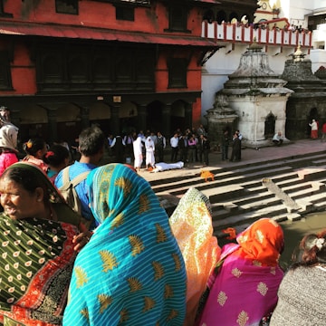 A group of people dressed in colorful traditional clothing gather near a stone platform by a body of water. The scene appears to be part of a cultural or religious ceremony with people observing solemnly. There are historic architectural structures in the background, adding to the setting's ambiance.