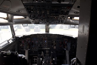 A cockpit with extensive control panels, gauges, and displays situated in the front of an aircraft. The view from the cockpit includes a partial outdoor scene with vehicles in the background.