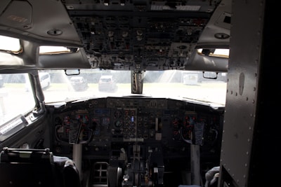 A cockpit with extensive control panels, gauges, and displays situated in the front of an aircraft. The view from the cockpit includes a partial outdoor scene with vehicles in the background.