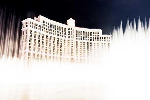 An elegant hotel building illuminated at night with a grand display of water fountains in the foreground. The structure features numerous windows and a rooftop dome, and the water jets create a dynamic visual effect against a dark sky.