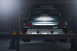 A car is elevated on a lift inside a dimly lit garage or workshop. The rear of the vehicle is visible, showcasing the metallic body and the word 'Carrera' on the back. The environment appears industrial with a focus on the car's detailing. The lighting casts a soft glow over the car, adding a dramatic effect.