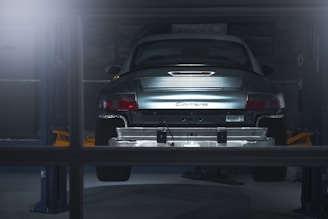 A car is elevated on a lift inside a dimly lit garage or workshop. The rear of the vehicle is visible, showcasing the metallic body and the word 'Carrera' on the back. The environment appears industrial with a focus on the car's detailing. The lighting casts a soft glow over the car, adding a dramatic effect.