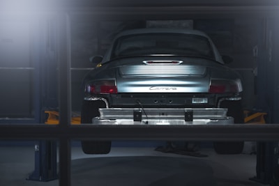 A car is elevated on a lift inside a dimly lit garage or workshop. The rear of the vehicle is visible, showcasing the metallic body and the word 'Carrera' on the back. The environment appears industrial with a focus on the car's detailing. The lighting casts a soft glow over the car, adding a dramatic effect.