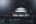 A car is elevated on a lift inside a dimly lit garage or workshop. The rear of the vehicle is visible, showcasing the metallic body and the word 'Carrera' on the back. The environment appears industrial with a focus on the car's detailing. The lighting casts a soft glow over the car, adding a dramatic effect.