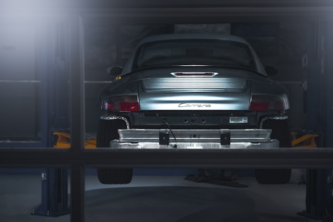 A car is elevated on a lift inside a dimly lit garage or workshop. The rear of the vehicle is visible, showcasing the metallic body and the word 'Carrera' on the back. The environment appears industrial with a focus on the car's detailing. The lighting casts a soft glow over the car, adding a dramatic effect.