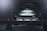 A car is elevated on a lift inside a dimly lit garage or workshop. The rear of the vehicle is visible, showcasing the metallic body and the word 'Carrera' on the back. The environment appears industrial with a focus on the car's detailing. The lighting casts a soft glow over the car, adding a dramatic effect.
