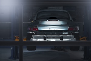 A car is elevated on a lift inside a dimly lit garage or workshop. The rear of the vehicle is visible, showcasing the metallic body and the word 'Carrera' on the back. The environment appears industrial with a focus on the car's detailing. The lighting casts a soft glow over the car, adding a dramatic effect.
