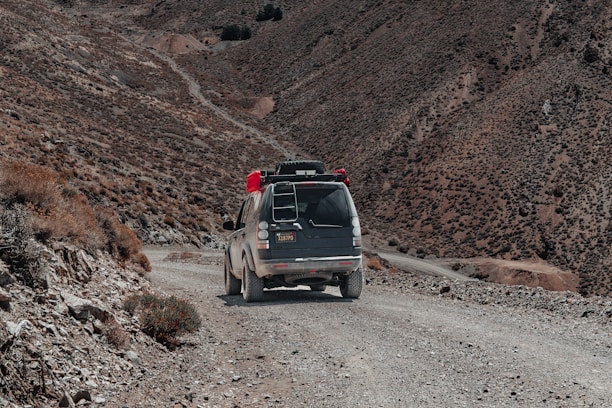 A rugged, dirt road winds through an arid mountainous landscape. A vehicle with gear and supplies on the roof rack travels down the path, surrounded by barren hills and rocky terrain.