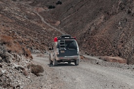 A rugged, dirt road winds through an arid mountainous landscape. A vehicle with gear and supplies on the roof rack travels down the path, surrounded by barren hills and rocky terrain.