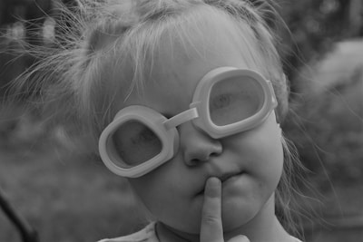 A black-and-white image of a young child wearing oversized goggles. The child has light hair and is making a thoughtful expression with one finger resting on their lips. The background is slightly blurred, suggesting an outdoor setting.