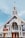 A large church with an A-frame structure and a prominent steeple topped with a cross. The building features tall white pillars at the entrance, wooden doors, and stained glass windows. Various banners with text are displayed on the exterior.