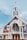 A large church with an A-frame structure and a prominent steeple topped with a cross. The building features tall white pillars at the entrance, wooden doors, and stained glass windows. Various banners with text are displayed on the exterior.