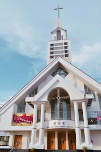 A large church with an A-frame structure and a prominent steeple topped with a cross. The building features tall white pillars at the entrance, wooden doors, and stained glass windows. Various banners with text are displayed on the exterior.