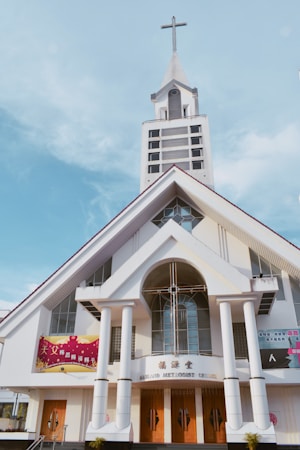 A large church with an A-frame structure and a prominent steeple topped with a cross. The building features tall white pillars at the entrance, wooden doors, and stained glass windows. Various banners with text are displayed on the exterior.