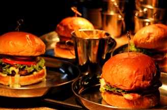 Four hamburgers are placed on metal trays, each containing lettuce, tomatoes, and sauce. A small bucket filled with french fries is also visible in the center, surrounded by the hamburgers. The setting appears to be dimly lit with a warm, inviting glow.