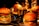 Four hamburgers are placed on metal trays, each containing lettuce, tomatoes, and sauce. A small bucket filled with french fries is also visible in the center, surrounded by the hamburgers. The setting appears to be dimly lit with a warm, inviting glow.