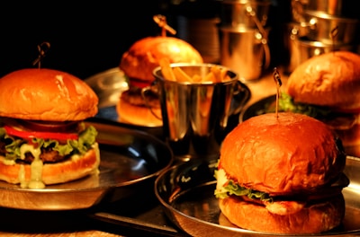 Four hamburgers are placed on metal trays, each containing lettuce, tomatoes, and sauce. A small bucket filled with french fries is also visible in the center, surrounded by the hamburgers. The setting appears to be dimly lit with a warm, inviting glow.