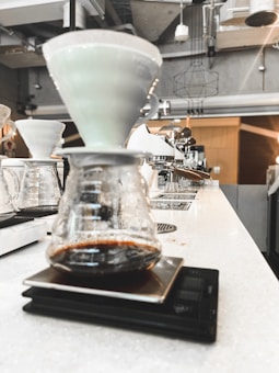 A close-up view of a pour-over coffee setup in a modern caf&eacute;. The image shows a glass carafe positioned on a digital scale with a ceramic dripper on top. The background features an industrial-style interior with visible light fixtures and coffee equipment.