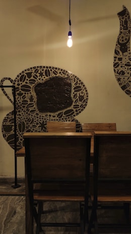 A cozy interior setting with a warm light bulb hanging from the ceiling. Wooden chairs are aligned next to a wall featuring artistic coffee cup-themed murals. The floor has a stone or concrete texture.