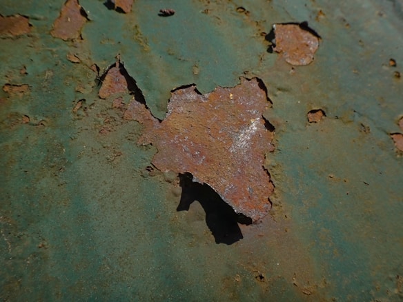 A close-up view of rust on a metal surface, with flaking and peeling paint. The rust appears in a variety of shades, contrasting with areas of remaining green paint. The surface texture is rough with jagged edges along the rust patches.