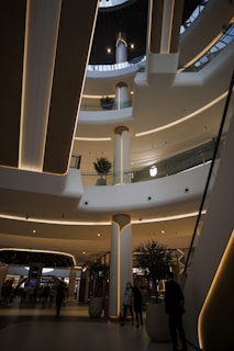 An indoor architectural scene of a modern shopping mall with multiple floors and a skylight at the top. The design features sleek, curved lines with embedded lighting. Several small trees in pots and people walking or standing in the space provide a sense of scale.
