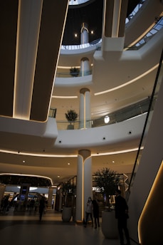 An indoor architectural scene of a modern shopping mall with multiple floors and a skylight at the top. The design features sleek, curved lines with embedded lighting. Several small trees in pots and people walking or standing in the space provide a sense of scale.