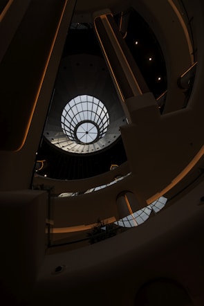 A modern architectural interior featuring a large circular skylight at the top, allowing natural light to stream in. The design includes multiple levels with sleek lines and a futuristic aesthetic. The structure appears symmetrical with metal and glass elements.