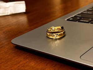 Two gold rings with embedded gemstones are placed on the edge of a laptop, which is set on a wooden surface. The light reflects off the rings, highlighting their shiny and intricate design.