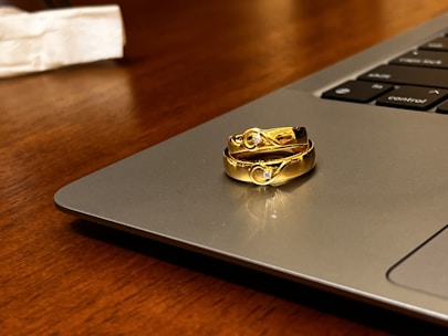 Two gold rings with embedded gemstones are placed on the edge of a laptop, which is set on a wooden surface. The light reflects off the rings, highlighting their shiny and intricate design.