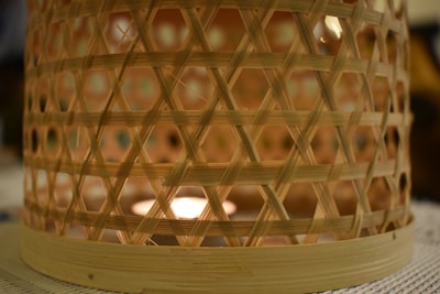 A woven bamboo basket that encases a source of light, creating a warm, glowing effect through its latticed structure. The intricate pattern of crisscrossed bamboo strips casts soft shadows on a textured surface.