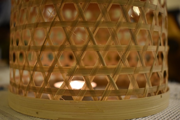 A woven bamboo basket that encases a source of light, creating a warm, glowing effect through its latticed structure. The intricate pattern of crisscrossed bamboo strips casts soft shadows on a textured surface.