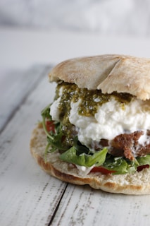 A close-up view of a sandwich featuring a variety of ingredients including leafy greens, white creamy cheese, crumbly brown patty, and green pesto sauce, all layered between soft, rustic bread.