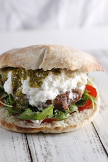 A close-up of a sandwich with a ciabatta bun, filled with arugula, tomatoes, a meat patty, white cheese, and a green sauce.