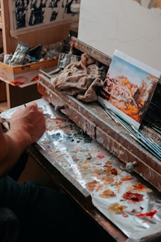 A workspace containing art supplies and a person's hand engaged in painting. The area is cluttered with paint tubes, brushes, and a cloth smudged with various paint colors. A printed image depicting an outdoor scene is propped against a wooden organizer, serving as a reference or inspiration.
