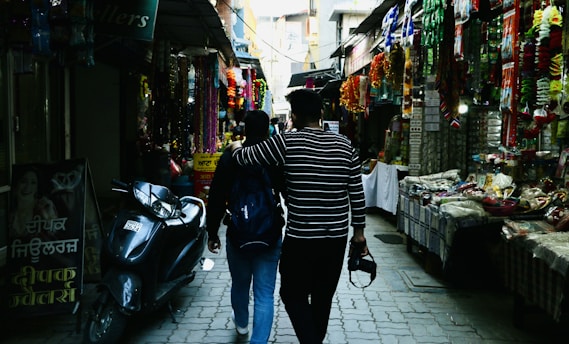 A narrow market street filled with colorful decorations and goods on display. Two people walk side by side, with one holding a camera, surrounded by vibrant hanging items and street vendors' stalls lining both sides. A parked scooter is visible on the left, next to a sign with text in a local script.
