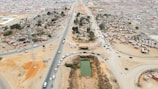 Aerial view of a developing urban area with a mix of paved and dirt roads. Dense clusters of small, shanty-like structures are situated on either side of a main road, with several vehicles traveling along the roads. There is a rectangular pond or water feature in the foreground and multiple trees scattered throughout the area.