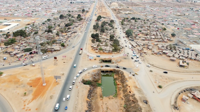 Aerial view of a developing urban area with a mix of paved and dirt roads. Dense clusters of small, shanty-like structures are situated on either side of a main road, with several vehicles traveling along the roads. There is a rectangular pond or water feature in the foreground and multiple trees scattered throughout the area.
