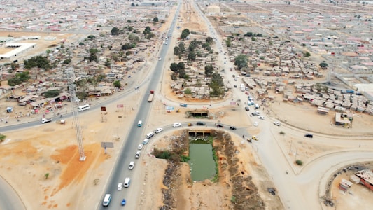 Aerial view of a developing urban area with a mix of paved and dirt roads. Dense clusters of small, shanty-like structures are situated on either side of a main road, with several vehicles traveling along the roads. There is a rectangular pond or water feature in the foreground and multiple trees scattered throughout the area.